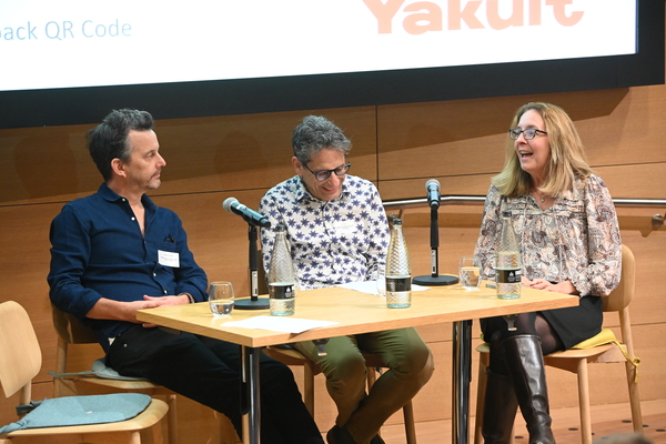 The three speakers are sitting at a table in an auditorium