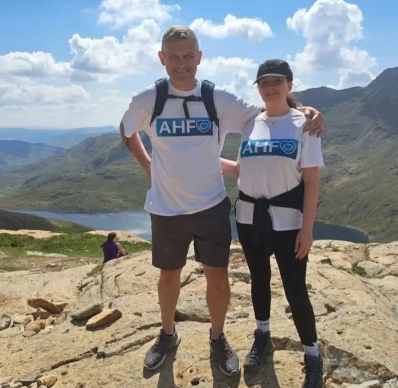 A man and woman stand on top of a mountain smiling in hiking gears wearing t-shirts that say 'An Hour for Others'