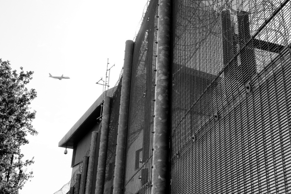 A black and white image of a plane flying over a fence with barbed wire