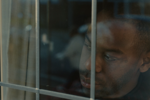 A Black man with a sullen expression looks through a window.