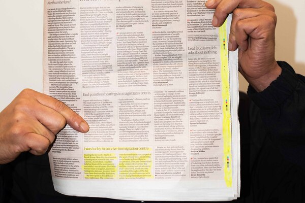 A person covers their face with a newspaper their hands are visible and one points to a letter about Brook House