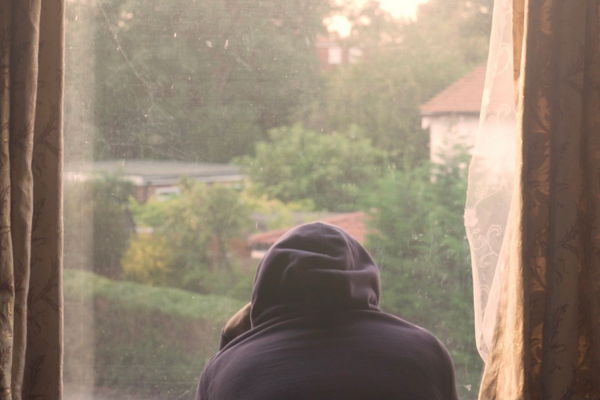 still from our film 'I need air'. A man is looking outside his window onto his garden with his back to us. He is wearing a dark hoodie.