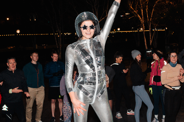freya, the human disco ball, wearing a silver disco suit and silver star shaped sunglasses, in front of a group of runners. 