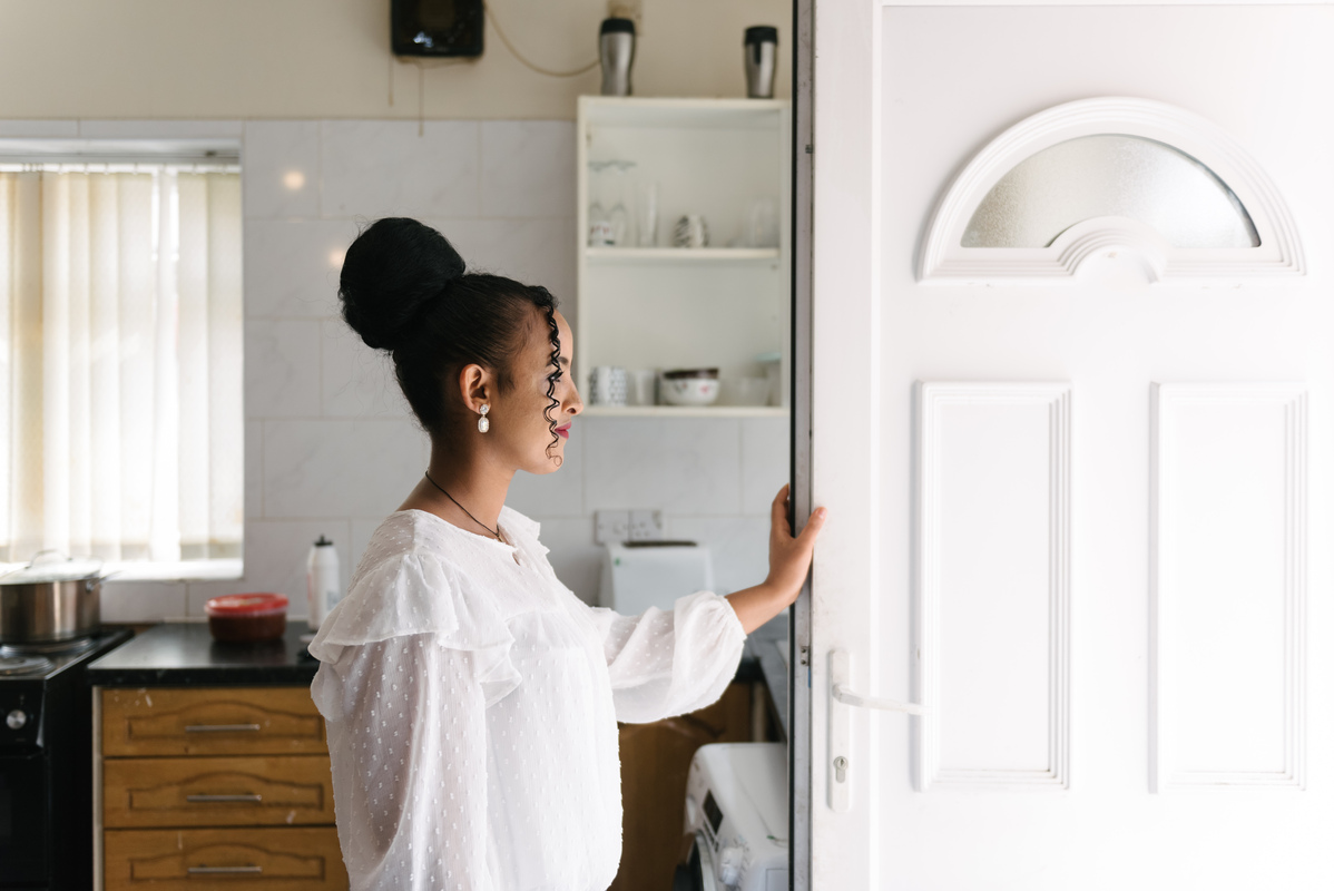 A Black woman stands inside her house, opening a door and smiling.