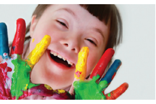 A girl with Down syndrome who is smiling with her hands held up covered in multi coloured paint
