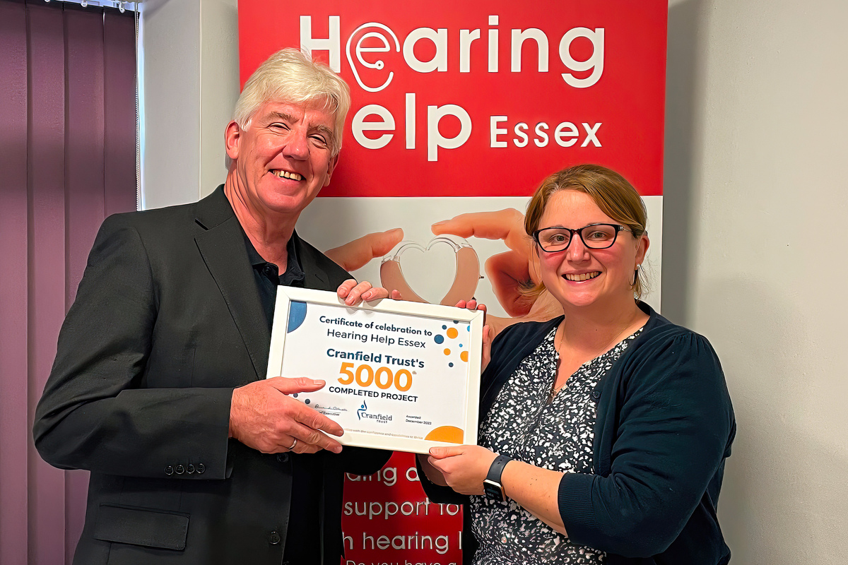 Sophie Ede, CEO of Hearing Help Essex receives a 5,000th project certificate from Chris Jullings, Cranfield Trust volunteer. Sophie and Chris are standing front of a banner with the Hearing Help Essex logo on a red background