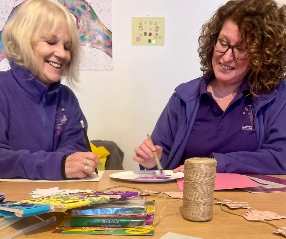 Two ladies sat next to each other, smiling, and doing craft activities. One lady had long curly brown hair and is wearing glasses, the other lady has blond shoulder length hair