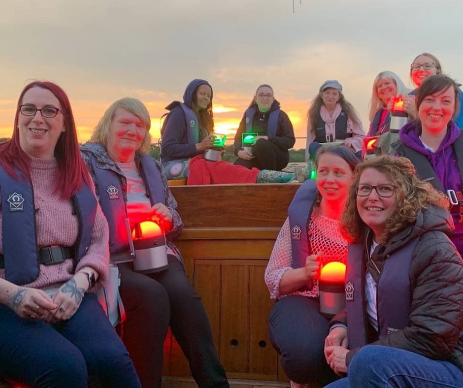 Photo of a big group of women sat in a horseshoe shape, holding candles at dusk, all smiling and looking towards the camera