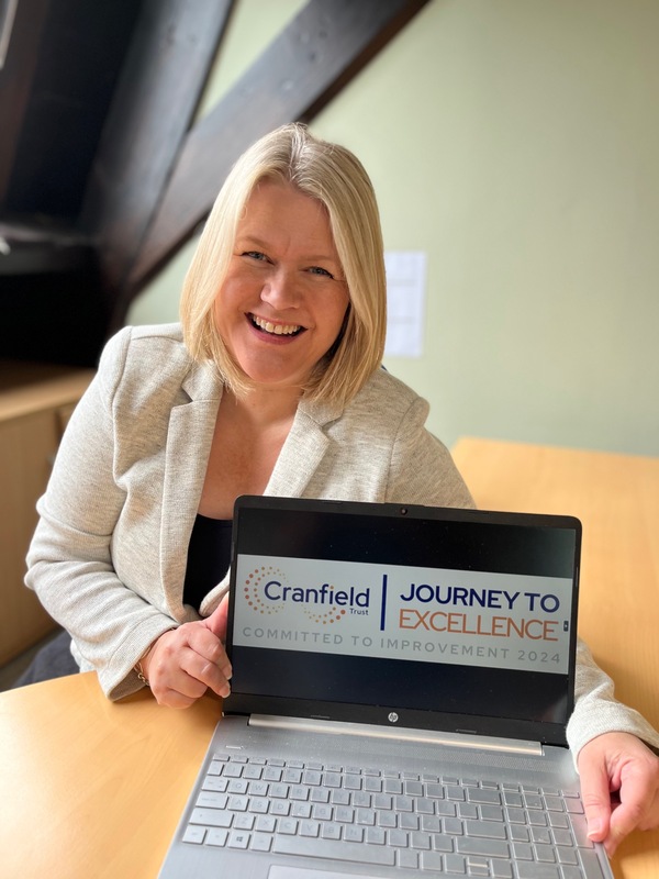 Photo of Building Circles CEO, Helen Kaye showing the new J2E Recognition Badge on her laptop. Helen had blond shoulder length hair and is wearing a cream jacket. She is smiling. 