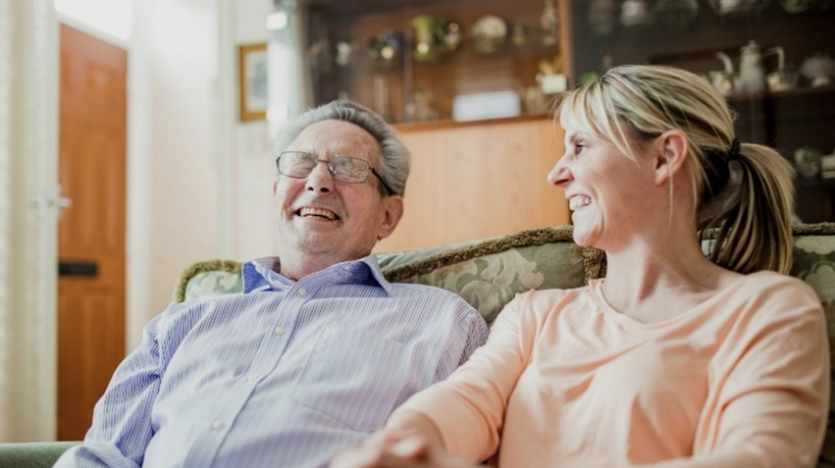 An older man and younger lady sit together on a sofa laughing and smiling.