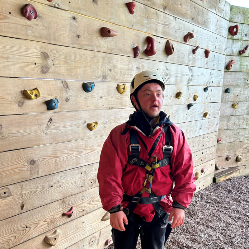 A man in climbing gear stands in front of a climbing wall.