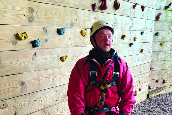 A young man with Down Syndrome wears protective clothing in front of a climbing wall