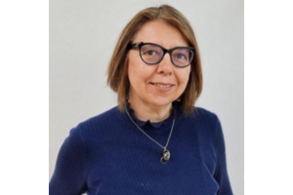 Photo of Alice Dabrowska, a female smiling. She is wearing glasses with a blue frame and has shoulder length brown hair. She is wearing a navy blue jumper and a necklace