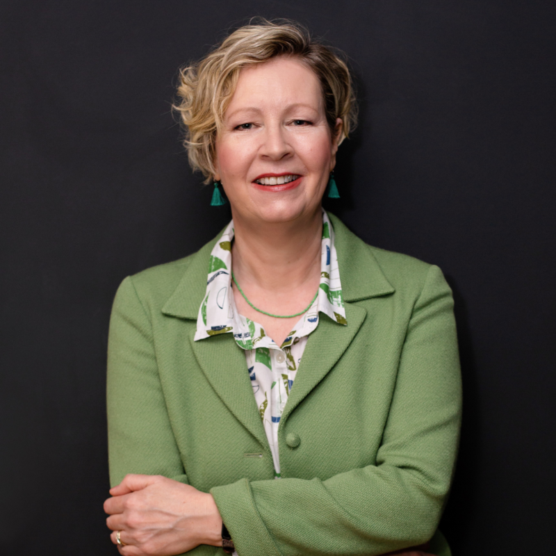 Head and shoulders photo of Emma. Emma is smiling and has her hands crossed and is wearing a green jacket with a white shirt underneath with green leaves on it the shirt. Emma has