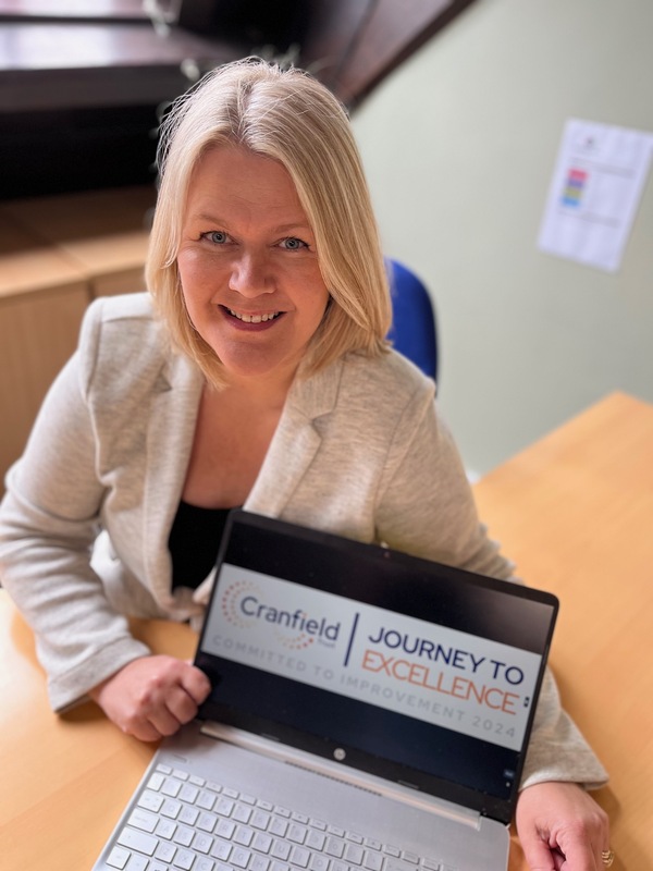 Head and shoulders image of Building Circles CEO, a lady with blond should length hair smiling and wearing a cream jacket. She is holding a laptop with the screen showing the J2E recognition badge 