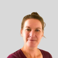 Head and shoulders photo of Ali Forbes. Ali is wearing a burgundy top with a round neck and is smiling at the camera. Ali has brown air up in bun