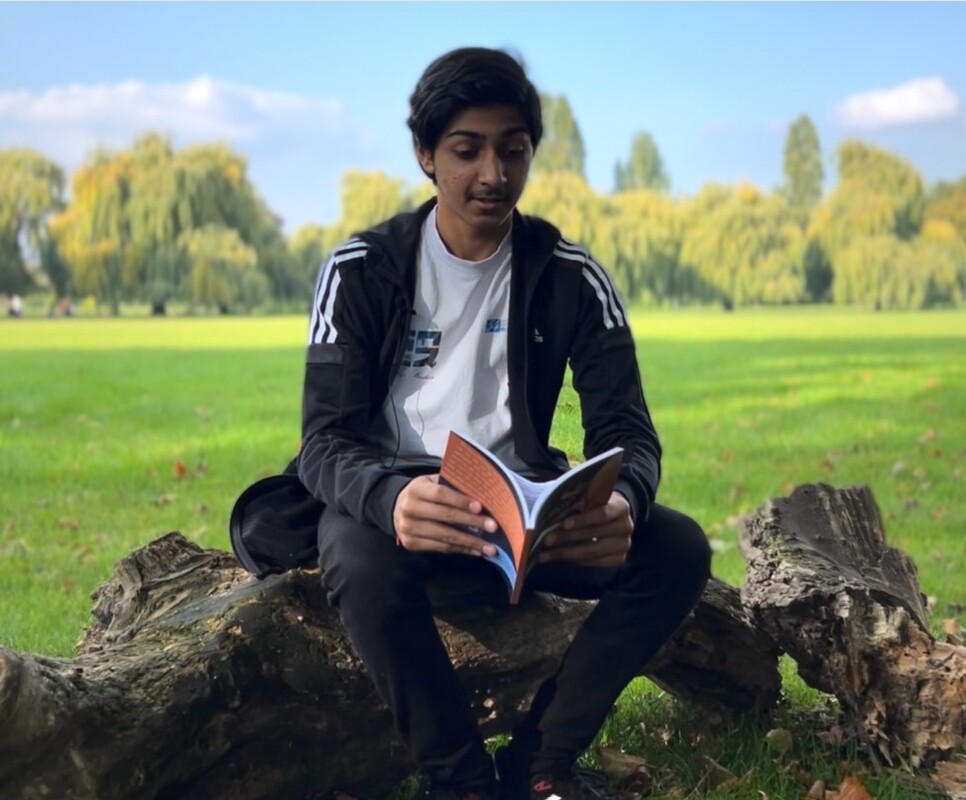 Photo of a young man reading a book sat on a big log in the park