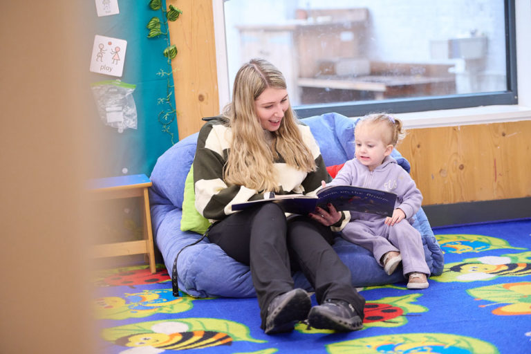 Photo of a young lady with long blond hair sitting on a bean bag with a little girl who is wearing purple and has blond hair in a bun, the young lady is reading a book to the young girl