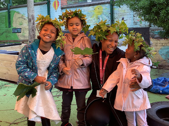Photo of three young girls dressed with wreaths on their heads, with their mum who is smiling at the smallest child