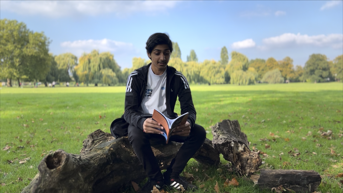 A young man sitting on a fallen tree trunk in a park, reading a book, with green open fields and trees in the background.