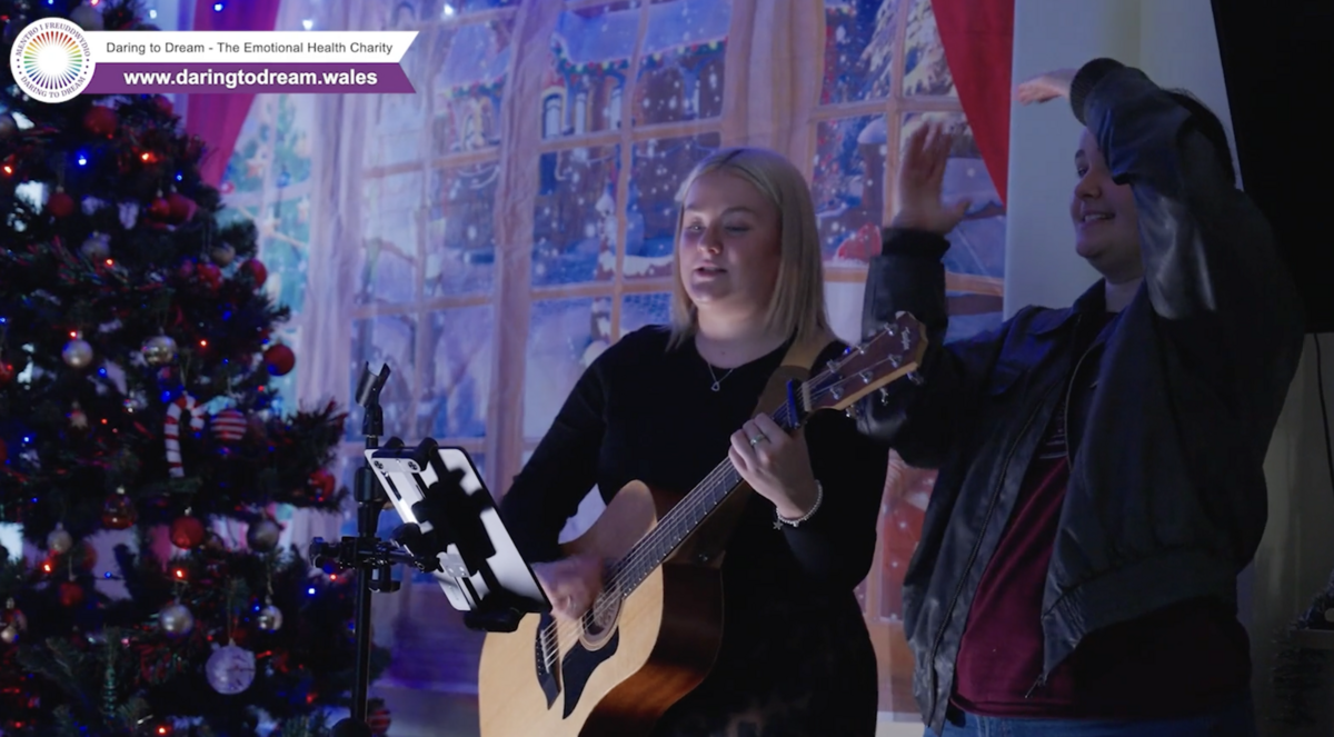 A young lady plays a guitar standing next to a Christmas Tree while a young man dances next to her.