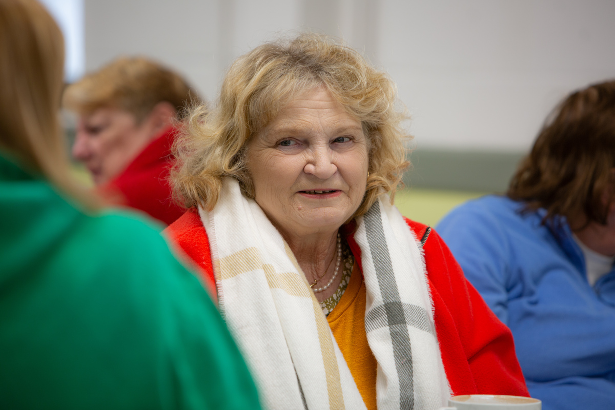 An older lady with blonde curly hair, wearing a red top and pale checked scarf is sat amongst a group of other ladies.