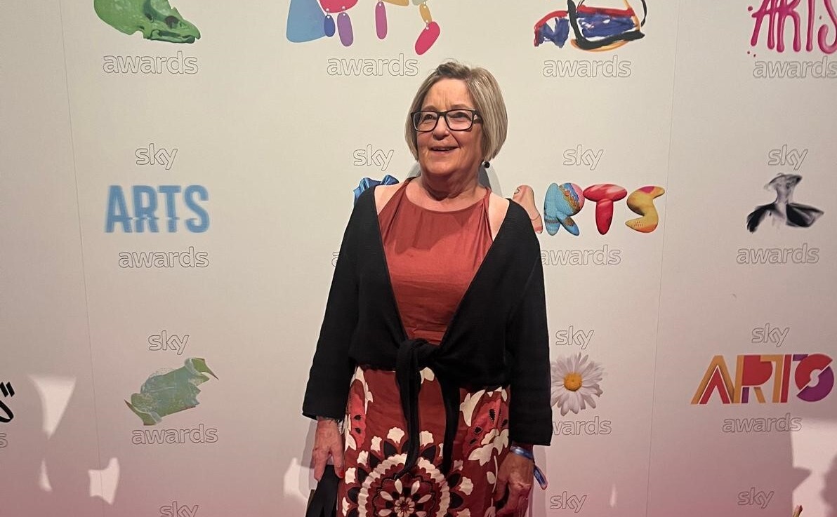 A woman with a blonde bob hair and glasses with a brown dress standing in front of an awards board