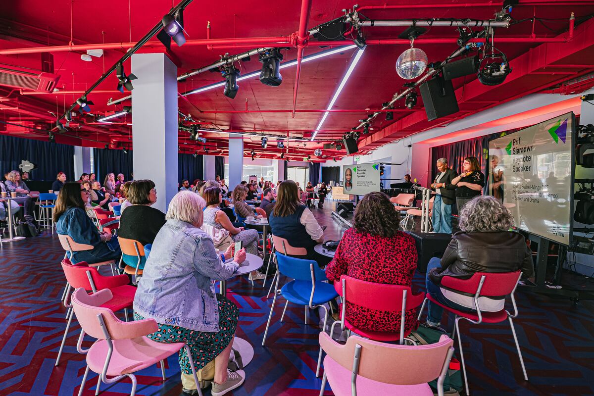 A room full of people watching a woman on the stage talking