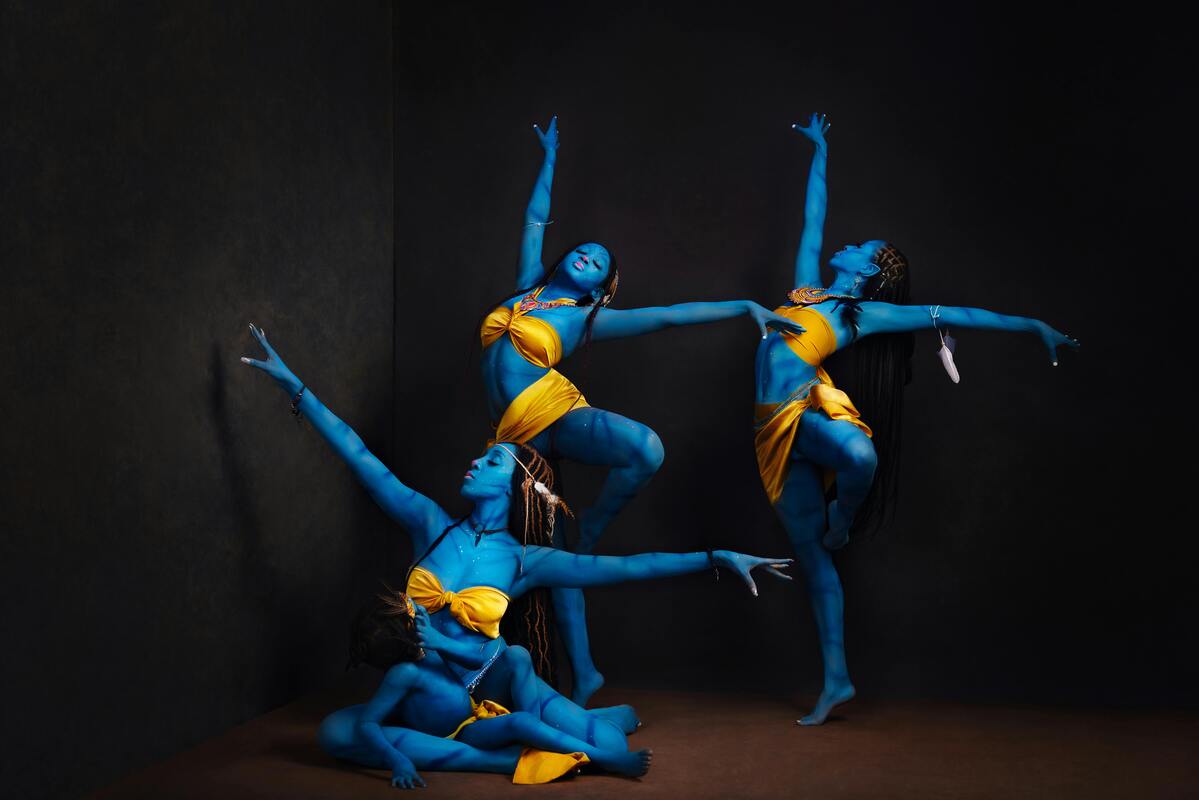 Three dancers with blue-painted skin and yellow costumes hold sculptural contemporary dance poses against a dark studio background.
