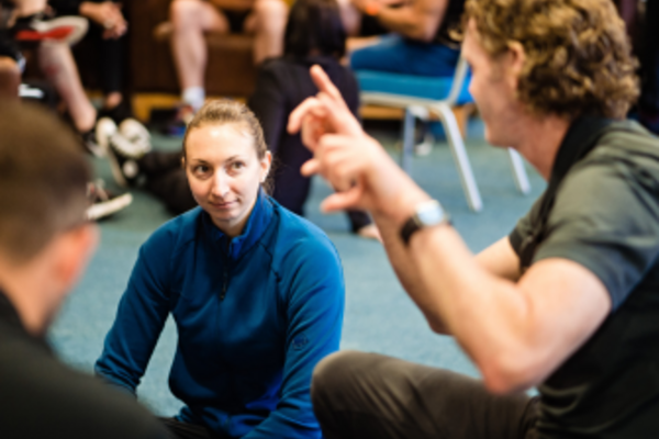 A woman wearing a blue jumper sitting with crossed legs on the floor listening as a man wearing a grey t-shirt speaks