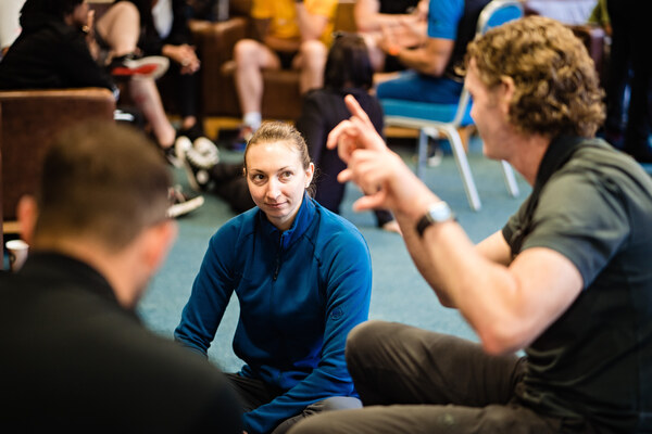 A man with animated hands talking to a woman, wearing a blue jumper, sitting on the floor listening.