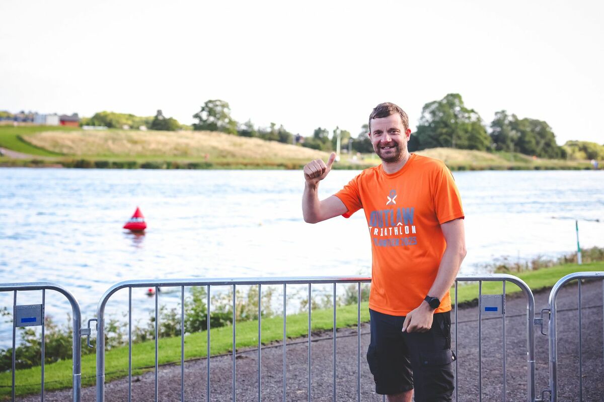 Liam Colebrook in an orange tshirt giving a thumbs up in front of a lake