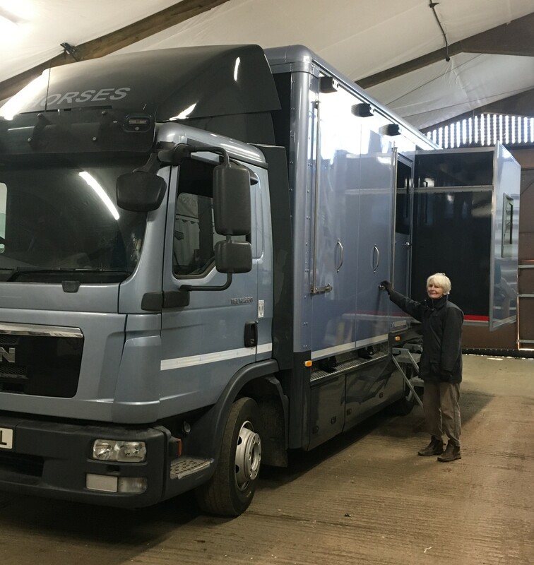 Picture of a large silver horsebox with a lady standing beside it