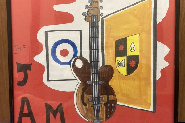 A framed artwork featuring a brown guitar in the center, with a red background. To the left of the guitar is the logo of "The Jam," a British mod band, with the word "The" written above and "JAM" written below in black letters. To the right of the guitar is a yellow and black shield-like emblem. Behind the guitar is a roundel design, with a blue circle around a red dot on a white background