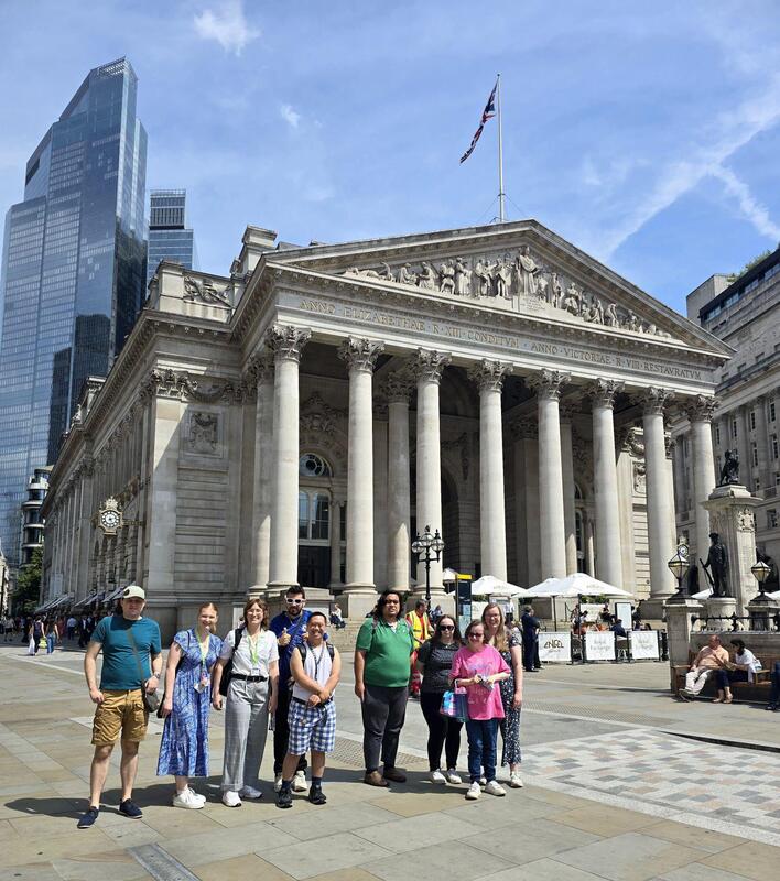 Group posing for a photo in front of the Royal Exchange building in London's financial district, with its distinctive neoclassical architecture featuring columns and pediment, and modern skyscrapers visible in the background under a partly cloudy sky.