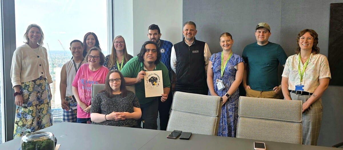 A group of twelve people pose together in a modern office meeting room with large windows showing a city skyline. One person in the front center holds up a colorful drawing that says "The Fidelis Partnerships." Everyone is smiling and dressed casually or in work attire, with some wearing lanyards. A small terrarium sits on the table in the foreground.
