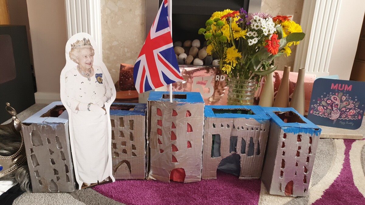 A miniature cardboard replica of Buckingham Palace with a small cardboard cut our of the Queen next to it 