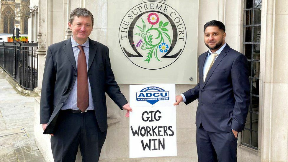 Two Uber Drivers hold up a sign saying Gig Workers Win outside the Supreme Court.