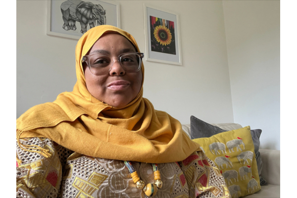 A photo of Annie Gibbs. Annie is a black woman wearing a patterned top, a yellow headscarf and glasses. She is sat on a couch with illustrations of an elephant and a sunflower behind them.