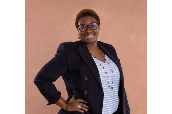 A photo of Olumide Adisa, orange background, with Olumide holding her hand on her hips. She is wearing glasses, a black blazer and a white stripey tshirt