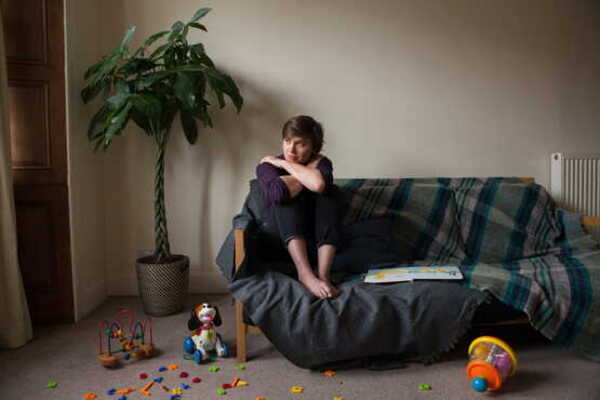 A photo of a woman sat on a sofa hugging her knees. She looks upset and children's toys are strewn around her.