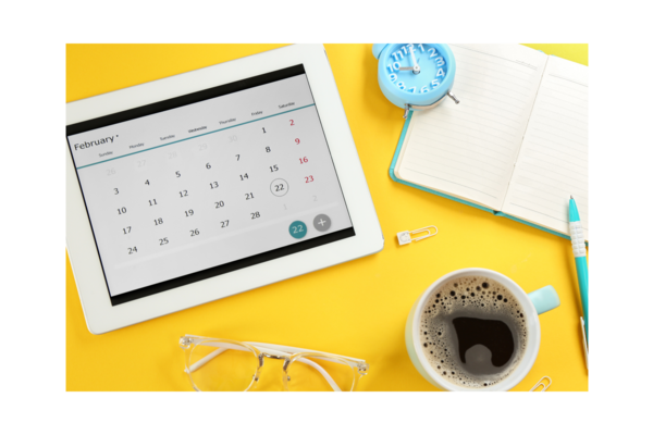 A photo of a yellow desk with a tablet showing a calendar, a cup of coffee, a pair of glasses, an alarm clock, a notepad and a pen.