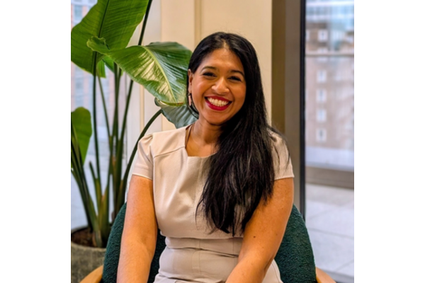 A photo of Rufsana Begum. She has long brown hair and is wearing smart clothes and smiling at the camera. Behind her is a plant and a window.