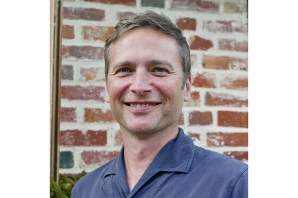 A photo of Jon Erin. He is standing in front of a brick wall, wearing a blue shirt and smiling at the camera.