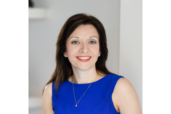 A photo of Esther Hamilton. She has brown hair and is wearing a blue dress and a gold necklace and is smiling at the camera.