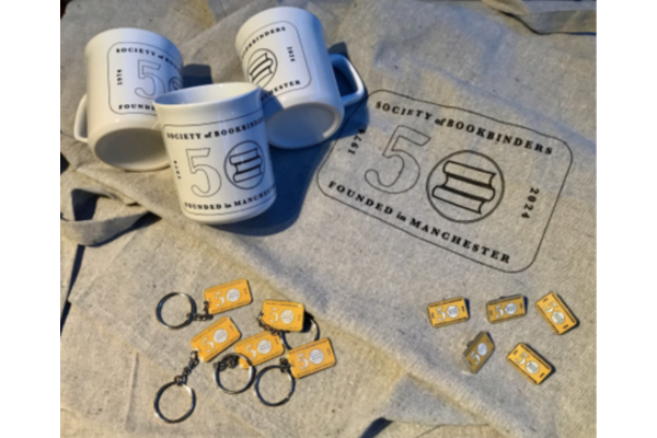 Photo of merchandise with the Society of Bookbinders fiftieth anniversary logo, including mugs, t-shirts, keychains, and badges