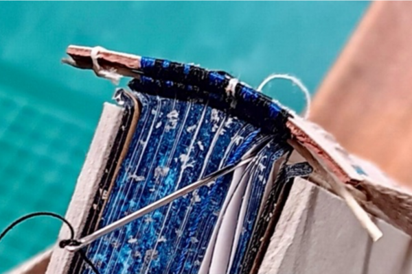 Photo of a headband being sewn with black and blue thread with a stripe of white, on a book with mottled blue edges speckled with silver