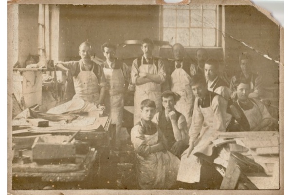 A sepia-toned or salt print photo of a group of binders wearing white aprons with tables covered in sheets of paper. The apprentices are at the front, one quite young.