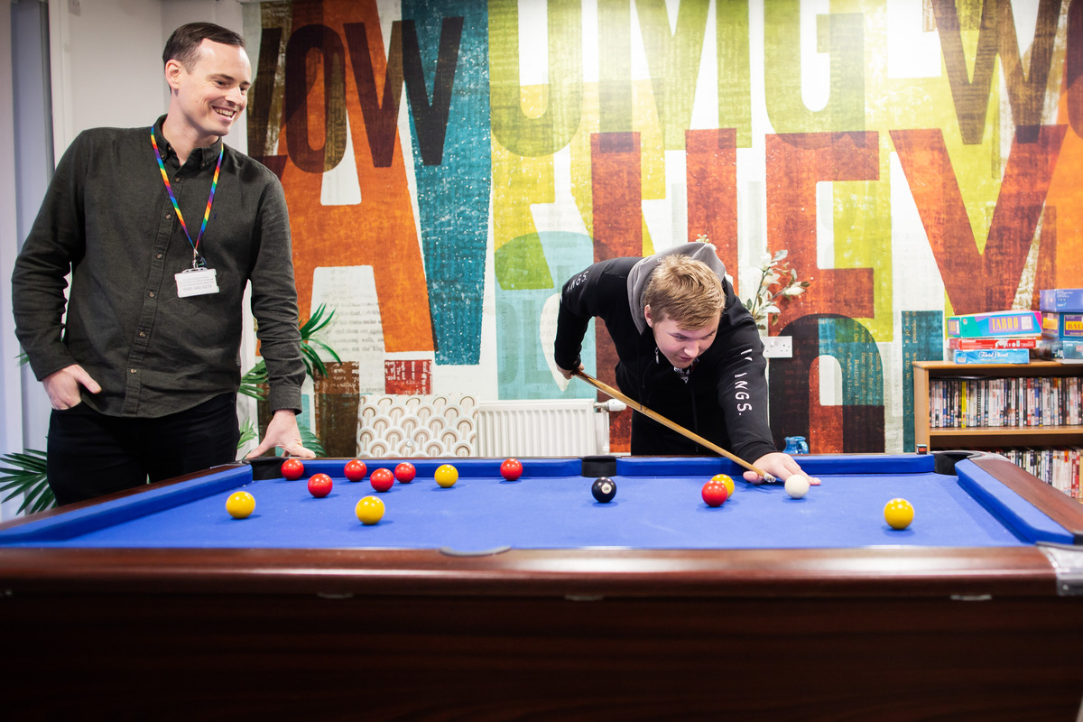 A young man is leaning over a pool table playing pool in the centre of the image and a male member of staff stands to the left watching and smiling, leaning a hand on the table. The background is a wall of brightly coloured letters spelling positive words.