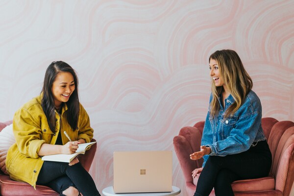 Two women sitting on pink suede chairs that have a shell like back. The young asian woman has a yellow funky jacket and a note pad and the young white women has her hand out. There is a laptop on a coffee table in front of them and they are smiling and laughing with each other
